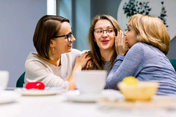 Drby s nejlepšími přáteli. Horizontální záběr dvou krásných žen šeptajících svým třetím přátelům do uší, zatímco popíjejí kávu s koláčky v kavárně. Ženy přátelé drbání během kávy. — Stock fotografie
