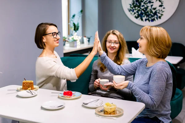 Üç kız arkadaş kafede kahve içip tatlı yiyorlar. Birbirlerine beşer beşer veren iki kadın, elinde bir fincan kahve ve gülümseyen üçüncü kadın.. — Stok fotoğraf