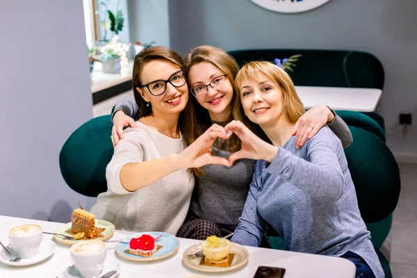 Hermosas tres mujeres jóvenes bebiendo una taza de café negro con deliciosos postres, sonriendo en el amor mostrando el símbolo del corazón y la forma con las manos. Concepto romántico . —  Fotos de Stock