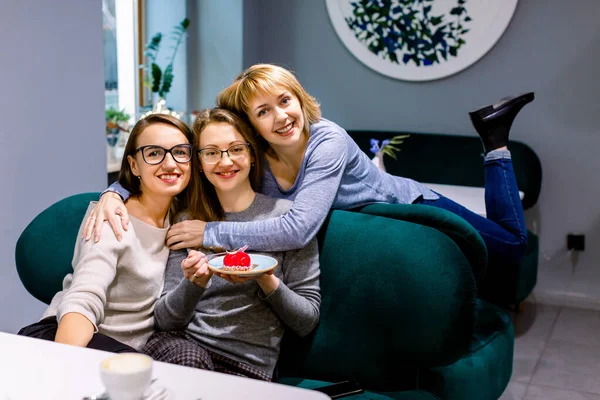 Vrouwen vrienden zitten op de bank in cafe, op zoek naar camera, een meisje houdt bord met heerlijke rode dessert — Stockfoto