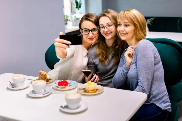 Tři mladé kamarádky - tři dívky, sedící u stolu v kavárně, klábosí, usmívají se, pijí kávu z bílých pohárů, jedí dezerty, jedí selfie. Životní styl, ležérní, radost z komunikace. — Stock fotografie