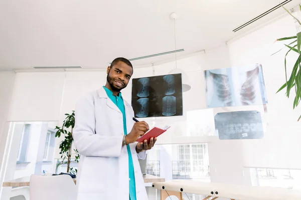Concetto di salute e medicina - sorridente medico africano che studia i raggi X dei pazienti mentre si trova in piedi nel moderno studio medico — Foto Stock