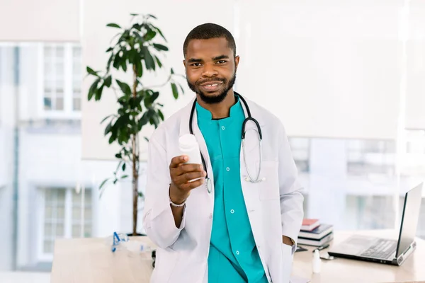 Medico africano maschio in uniforme medica e cappotto bianco, sorridente, bottiglia con pillole o capsule, in piedi in ufficio ospedaliero — Foto Stock