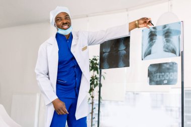 Tıbbi üniformalı ve maskeli genç Afro-Amerikan erkek doktor hastaların göğüs röntgenlerini inceliyor.