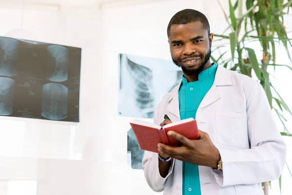 Portrait d'un jeune homme africain médecin en costume médical, regardant la caméra, tenant un otebook rouge et un stylo, debout dans un bureau moderne avec une radiographie sur le fond — Photo