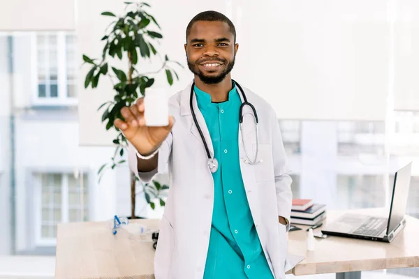 Giovane Dottore Africano Maschio con Stetoscopio e in mantello medico Tenere farmaci per i pazienti in trattamento, sorridente e in posa in clinica moderna. Focus sul viso — Foto Stock