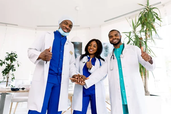 Doctors team. Health care. Three happy African doctors stack hands together as team for motivation, thumbs up