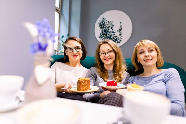 Women friends looking at camera, smilikng, holding cakes, three girl friends drinking coffee in the cafe and eating desserts, women during lunch time in cafe