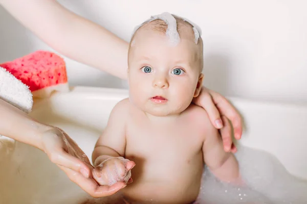 Glücklich lachendes Baby, das ein Bad nimmt und mit Schaumblasen spielt. Kind in der Badewanne. Säuglingswäsche und Baden. Unerkennbare Mutter badet ihr Baby in weiße kleine Plastikfledermaus. — Stockfoto