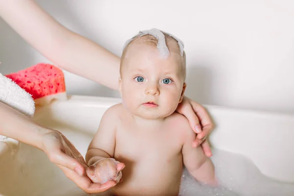 Happy laughing baby taking a bath playing with foam bubbles. Little child in a bathtub. Infant washing and bathing. Unrecognizable mother bathing her baby in white small plastic bat. — Zdjęcie stockowe