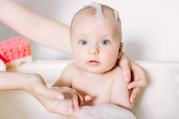 Glücklich lachendes Baby, das ein Bad nimmt und mit Schaumblasen spielt. Kind in der Badewanne. Säuglingswäsche und Baden. Unerkennbare Mutter badet ihr Baby in weiße kleine Plastikfledermaus. — Stockfoto