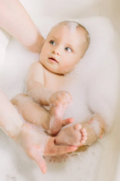 Happy laughing baby taking a bath playing with foam bubbles. Little child in a bathtub. Infant washing and bathing. Baby looking upwards in a plastic tub full of foam. — Stock Photo, Image
