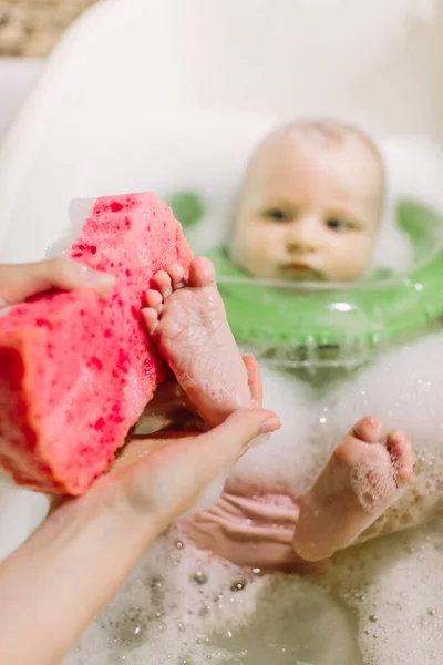 Μωρό κολύμβηση με δαχτυλίδι πράσινο λαιμό κολύμπι. μάνα τρίβει τα πόδια του μωρού με ένα κόκκινο σφουγγάρι — Φωτογραφία Αρχείου