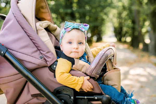 Ευτυχισμένο κορίτσι σε καροτσάκι μωρού που παίζει σε καροτσάκι με φόντο τη φύση. Πορτρέτο χαριτωμένο μικρό όμορφο κορίτσι 9 μηνών κάθεται ή καροτσάκι και περιμένει τη μαμά. — Φωτογραφία Αρχείου