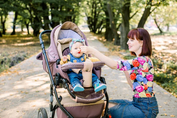 Η μαμά ταΐζει το παιδί περπατώντας με καροτσάκι στο πάρκο. . — Φωτογραφία Αρχείου