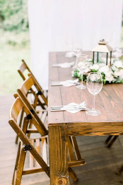 Ślub w stylu rustykalnym. Dekoracja stołu weselnego. Drewniany stół ozdobiony nożami i widelcami, kieliszki do wina, zieleń i kwiaty złotą latarnią — Zdjęcie stockowe