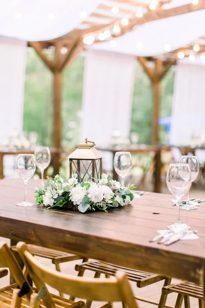 Hochzeitsdekorationen im rustikalen Stil auf Holztisch, Blumengesteck und goldener Laterne. Tisch mit Besteck und Geschirr, Weingläser. — Stockfoto