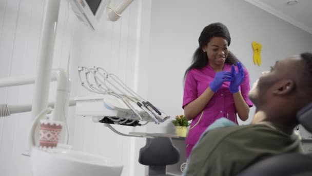 Joven dentista africana vestida con un traje rosa que se prepara para un examen de paciente, con guantes médicos azules. Hombre africano guapo paciente — Vídeos de Stock