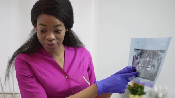 Una joven y bonita dentista africana vestida con un traje médico rosa y guantes azules muestra a la paciente una radiografía dental panorámica de las mandíbulas, y explica el tratamiento. Clínica dental, concepto de cuidado bucal — Vídeos de Stock