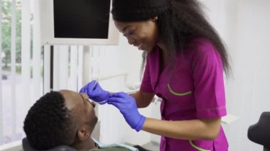 Diş ve sağlık sigortası konsepti. Profesyonel gülümseyen Afro-Amerikan kadın dişçi pembe tıbbi takım elbiseli ve mavi eldivenli modern diş sağlığı kliniğinde genç Afrikalı erkek hastaların dişlerini inceliyor.