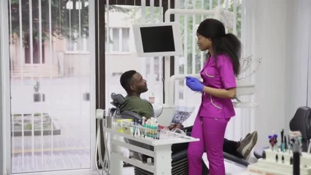 Zubní koncept, zdravotní péče, ústní péče. Mladá krásná africká žena zubař diskutuje o strategii léčby s pacientem, africký muž, sedí v křesle a drží xray image čelistí — Stock video