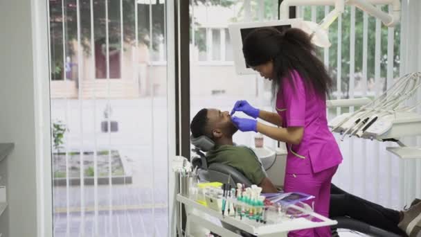 Odontóloga africana bastante joven examinando a su guapo paciente masculino de piel oscura con herramientas dentales profesionales en la clínica moderna. Concepto de odontología y cuidado bucal — Vídeo de stock