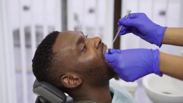 在现代牙科诊所，专业女牙医手拉手，戴蓝色防护手套，为年轻而英俊的非洲男性患者治疗牙 — 图库视频影像