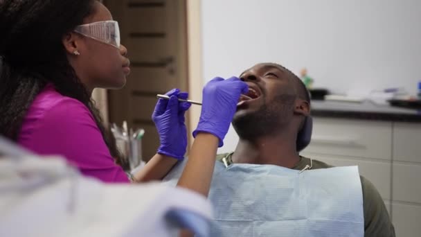 Conceito odontológico e de saúde. Dentista mulher africana profissional em terno médico rosa, óculos de proteção e luvas azuis examinando dentes de jovem paciente masculino africano, na clínica odontológica moderna — Vídeo de Stock