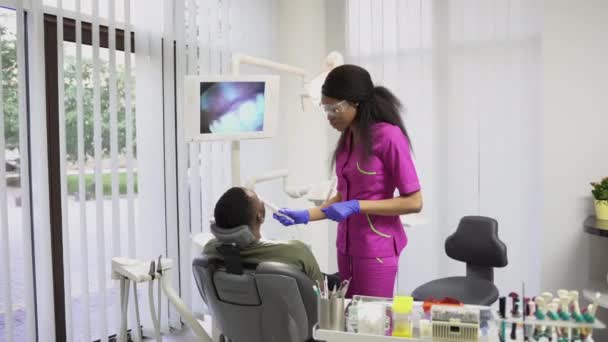 Стоматология, здравоохранение, пероральная и зубочистка. Молодая красивая африканская женщина-стоматолог проводит диагностику с помощью стоматологической визуографии и беседует с пациентом, африканцем, сидящим в кресле — стоковое видео