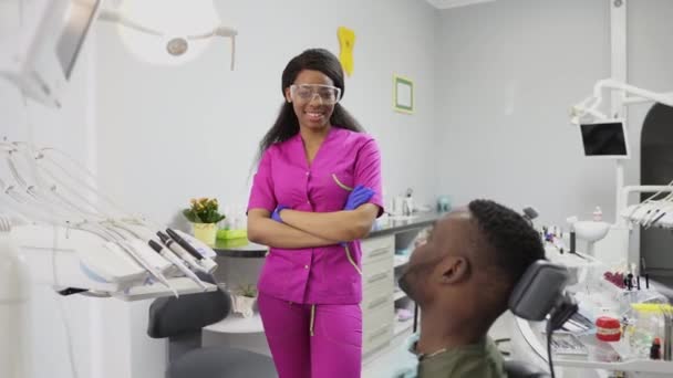 Bastante africana dentista feminina em terno rosa, óculos de proteção e luvas de látex azul posando para câmera, sorrindo, mostrando OK sinal e polegar para cima. Paciente africano do sexo masculino sentado na cadeira dentária — Vídeo de Stock