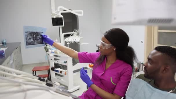 Joven y hermosa dentista africana hablando con su guapo paciente masculino de piel oscura en la clínica dental, mostrando una imagen de rayos X de sus dientes. Concepto de cuidado dental y oral — Vídeo de stock