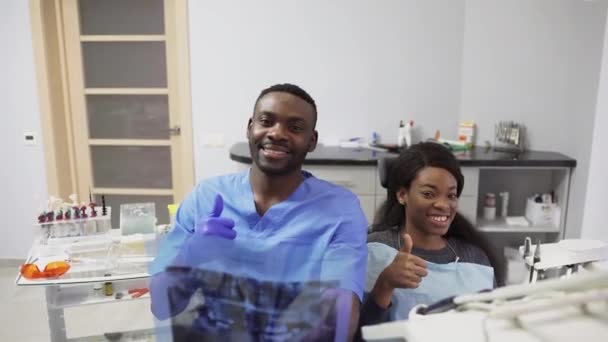 牙科诊所的概念，医疗保健，口腔和牙齿护理。年轻的非洲男人牙医拿着X光的牙齿形象和漂亮的非洲女病人的前视图，他们举起大姆指，微笑着 — 图库视频影像