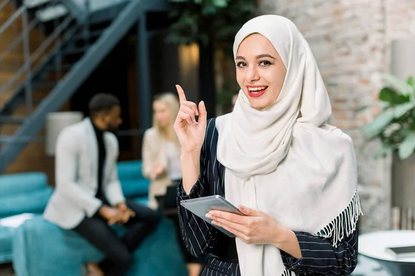 흰색 히잡을 쓴 예쁘고 젊은 무슬림 사업가가 현대의 다락방 사무실에서 서 디지탈 태블릿 PC 를 들고 손가락을 들고 웃으면서 비즈니스 편의점 배경에 서서 — 스톡 사진