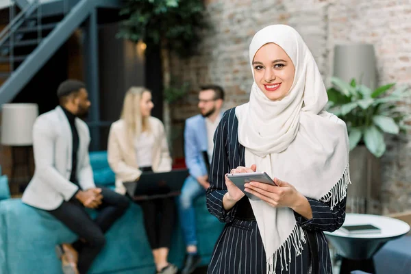 De cerca la cara de retrato de la atractiva mujer musulmana en hijab blanco, sosteniendo la tableta digital, mirando a la cámara y sonriendo, de pie en sus colegas de oficina multiétnicos fondo — Foto de Stock