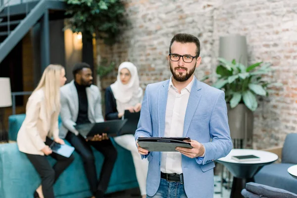 Porträt eines professionellen gutaussehenden kaukasischen Geschäftsmannes mit Brille, der einen digitalen Tablet-Computer vor dem Hintergrund seiner multiethnischen Kollegen im modernen Büro hält — Stockfoto