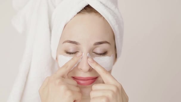 Porträtt av vacker vit ung kvinna med hår insvept i vit handduk, applicera ansiktslera mask röra hennes ansikte. Skönhetsbehandlingar. Dagspa. — Stockvideo