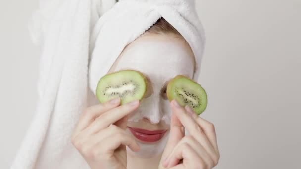 Close up de uma menina com máscara branca no rosto e uma toalha na cabeça fechando os olhos com fatias de frutas kiwi e sorrindo. Conceito de cuidados com a pele facial — Vídeo de Stock