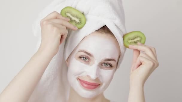 Young woman with white mud mask on face, on white background, posing with kiwi slices and smiling looking at camera. Teen girl taking care of skin, purifying the pores. Beauty treatment. Skincare. — Stock Video