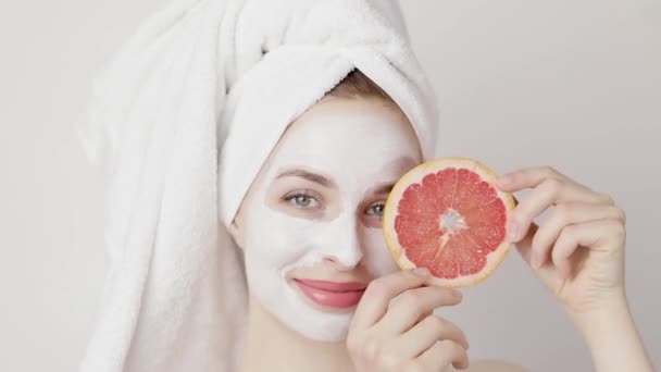 Wanita Spa dengan masker wajah. Wanita cantik dengan masker wajah lembab di wajahnya memegang sepotong jeruk menyembunyikan satu mata dan tersenyum. Pengobatan wajah. Kosmetologi, keindahan dan spa — Stok Video