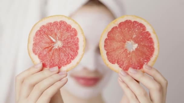 Smiling young cute girl with a white towel on her head and white nutritious mask on her face hiding her eyes with two slices of grapefruit and posing to camera — Stock Video