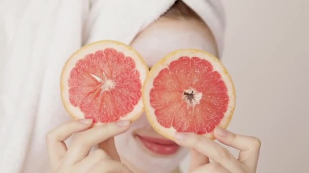 Zâmbind tânără fată drăguță cu un prosop alb pe cap și o mască nutritivă albă pe fața ei ascunzându-și ochii cu două felii de grapefruit și pozând la cameră — Videoclip de stoc