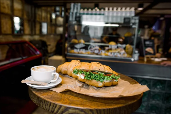 Šálek kávy cappuccino a lahodný francouzský domácí croissant s čerstvým salátem a zeleninou na dřevěném kulatém stole v ranní kavárně City, rozmazaný kavárna interiér na pozadí — Stock fotografie