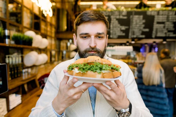 Have a break, lunch time. Close up of a cheerful handsome young bearded man, holding plate with fresh croissant and enjoying his break while smelling his favourite food, sitting in cafe