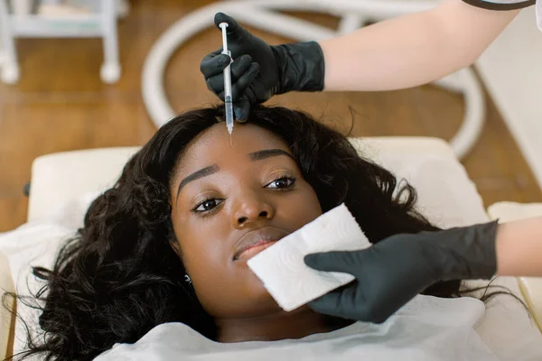 Aesthetic surgery. Young African Woman having injection on forehead in salon, top angle view. Pretty young doctor cosmetologist in black rubber gloves making an injection into her forehead skin