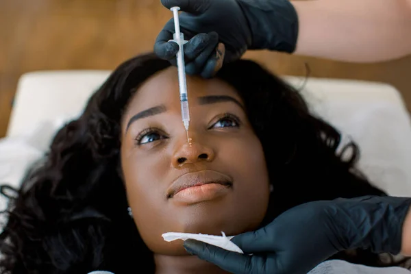 Закріпіть увагу на руки лікаря - косметолога, який робить реабілітаційну терапію, робить ін "єкції на обличчі для затягування і згладжування шкіри прекрасної африканської жінки в клініці. — стокове фото