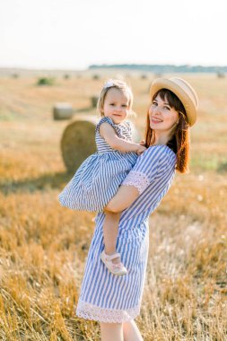Hasır şapkalı, çizgili elbiseli, yaz buğday tarlasında arka planda saman balyalarıyla gevşeyen sarışın kızını gün batımında kucağında tutan oldukça koyu renk saçlı bir kadın. Kırsalda yürü