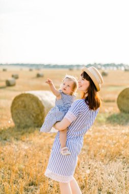 Hasır şapkalı ve çizgili elbiseli güzel bir anne ellerinde küçük tatlı bebeği gökyüzüne bakıyor, arka planda buğday saman balyalarıyla yaz tarlasında yürüyor