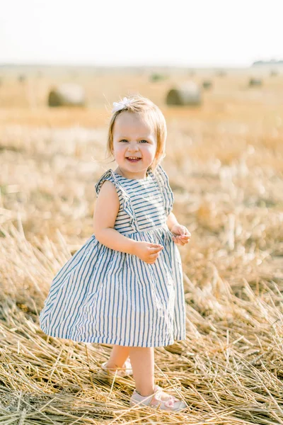 Χαριτωμένο μικρό κοριτσάκι σε κομψό ριγέ φόρεμα στέκεται στο παρασκήνιο του ηλιόλουστο σιτάρι καλοκαιρινό πεδίο με σανό στοίβες ή μπάλες, θέτοντας στην κάμερα και χαμογελώντας — Φωτογραφία Αρχείου