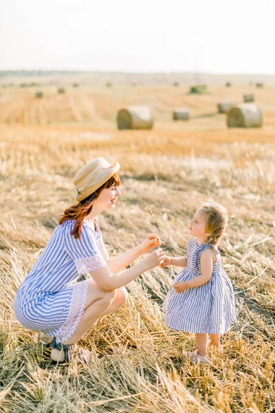 Tiro ao ar livre de mulher bonita com sua pequena filha criança vestindo vestidos listrados se divertindo no campo de trigo com pilhas de feno no dia de verão, brincando com espiguetas de trigo — Fotografia de Stock