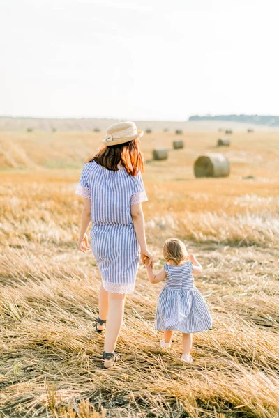 Mutlu anne ve 2 yaşında benzer çizgili mavi elbiseler giyen küçük bir kızın yaz portresi. Güneşli bir günde arka planda saman balyalarıyla el ele yürüyorlar. Arka plan — Stok fotoğraf